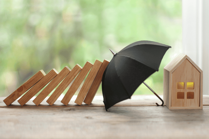 A toy wooden house is protected by a black umbrella, which prevents a line of dominoes (mid-fall) from hitting the house.