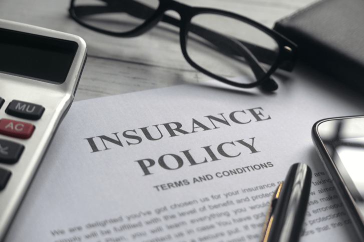 A close-up image of an anonymised insurance policy laid out on a desk, surrounded by a calculator, a pair of glasses, and a pen